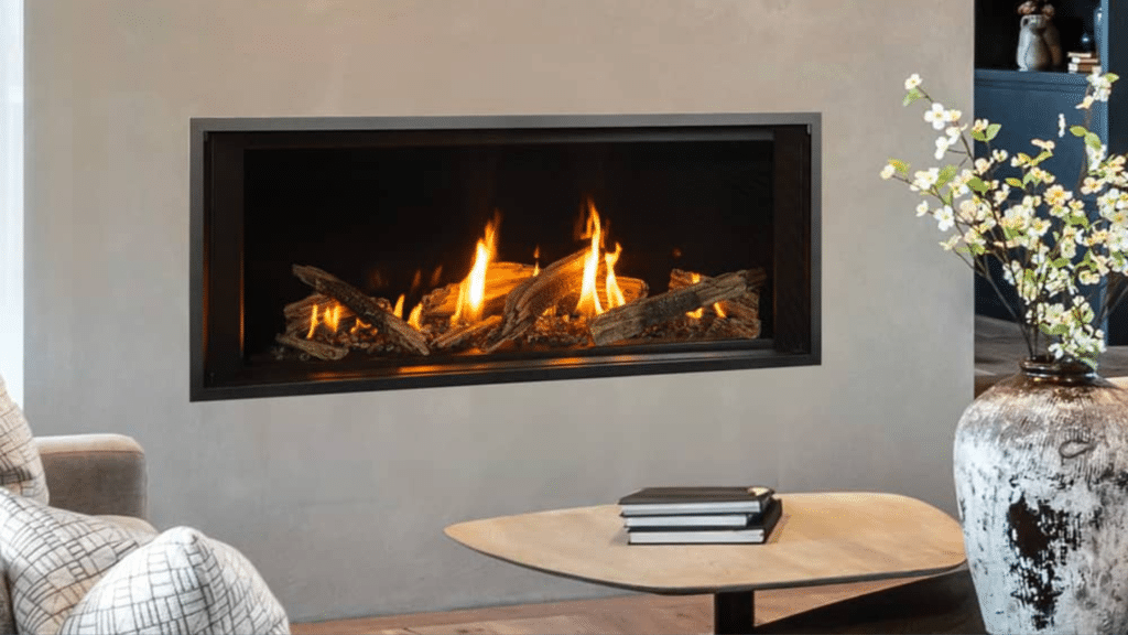 Modern gas fireplace with a glass front, burning logs inside. A vase with flowers and stacked books on a wooden table nearby.